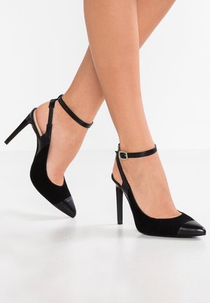 Voeten met zwarte slingback hoge hakken met enkelbandjes en puntige neuzen, geposeerd tegen een witte achtergrond.