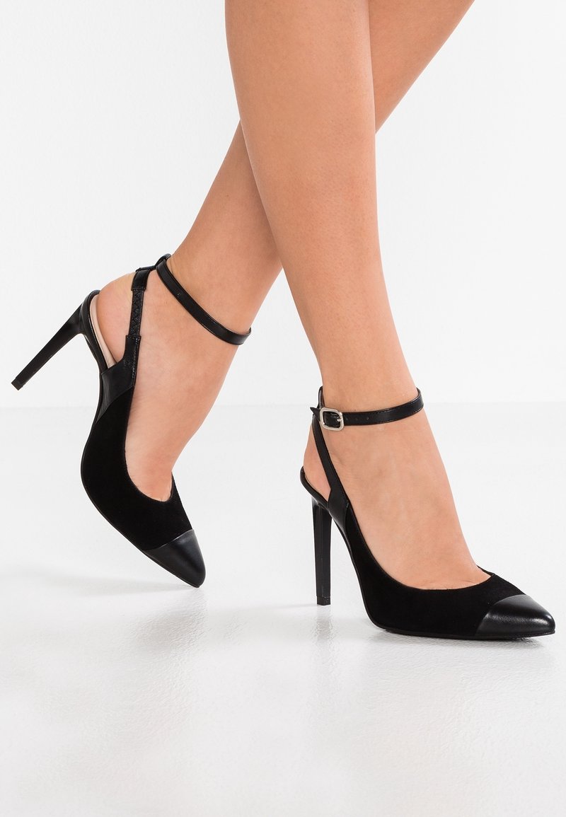 Pieds portant des chaussures slingback noires à talons hauts avec des lanières à la cheville et des bouts pointus, posés sur un fond blanc.