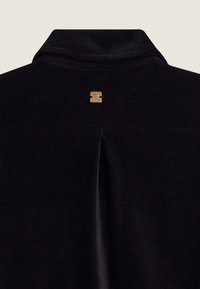 Tessuto di velluto a coste nero con una sottile trama a coste. Presenta una piega sul retro e un piccolo logo dorato vicino al colletto.
