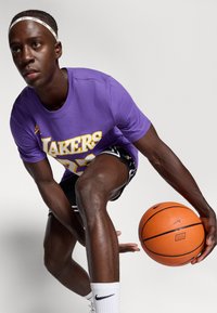 Ljubičasta majica Los Angeles Lakers sa bijelim i žutim natpisom, crne kratke hlače, drži narančastu košarkašku loptu. Bijele Nike čarape i traka za glavu. Neutralna pozadina.