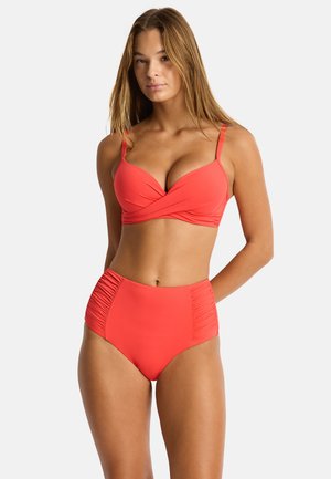 Młoda kobieta ubrana w koralowy bikini z wysokim stanem i marszczonymi bokami, stoi z rękami za plecami na jednolitym białym tle.