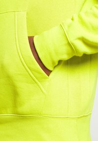 En mörkhyad hand inskjuten i framfickan på en klar gulgrön hoodie med synliga söm detaljer.