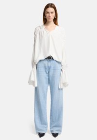 Blouse blanche avec des manches volumineuses, présentant des détails froncés aux épaules, portée avec un jean large bleu clair et des escarpins noirs à bout pointu.