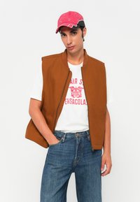 Jeune homme portant une casquette rouge, une veste sans manches marron, un t-shirt graphique blanc et un jean bleu, avec une main dans sa poche, sur un fond uni.