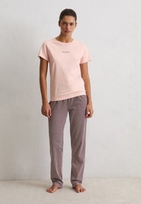 Vrouw staat blootsvoets tegen een effen muur, gekleed in een lichtroze T-shirt met korte mouwen en een losse broek met een patroon, kijkt recht vooruit met een neutrale uitdrukking.