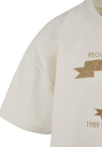Kreemist puuvillast t-särk lühikeste varrukatega. Kujundusel on kuldne broderie ja bänner, millel on tekst "REGISTREERITUD" ja "1989".