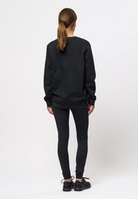 Personne aux cheveux bruns attachés en queue de cheval, portant un sweat-shirt noir, des leggings noirs et des baskets noires, debout de dos sur un fond clair uni.