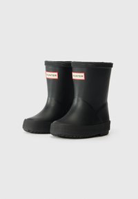 Bottes de cheville en caoutchouc noir avec une semelle texturée, ornées d'étiquettes "HUNTER" rouges et blanches à l'avant et d'un design lisse et arrondi.