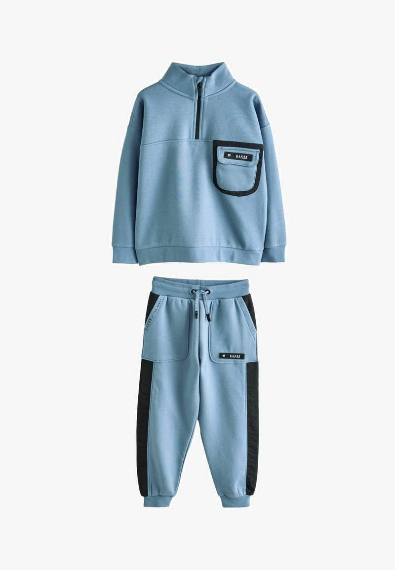 Svetlo modri trenirka iz mehkega blaga, ki ima zgornji del s polovičnim zadrgo in žepom na prsih ter ustrezne hlače z črnimi bočnimi paneli.