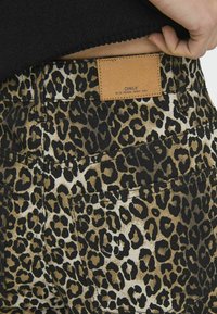 Hlače s uzorkom leoparda koje imaju crno-smeđu paletu boja, kožnu oznaku na leđima i vidljive detalje šivanja.