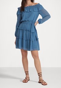 Kvinna som bär en off-shoulder klänning med blåa prickar, rufsar och midjeband, i kombination med bruna snörade platta sandaler.