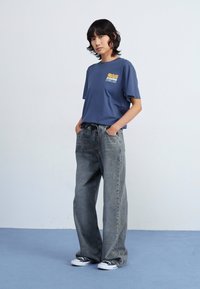 Blå T-skjorte med kort erm og grafikk på brystet, kombinert med løse grå jeans og svart-hvite joggesko. Enkelt, avslappet snitt.
