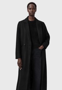 Abrigo largo de lana negra con cuello en pico, cierre de un solo botón y bolsillos laterales, combinado con un suéter negro texturizado y jeans oscuros.