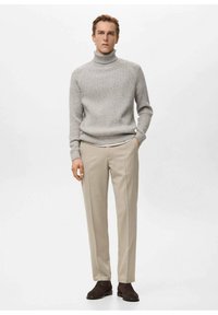 Man draagt een lichtgrijze geribbelde coltrui, beige op maat gemaakte broek en donkerbruine nette schoenen, staand tegen een witte achtergrond.