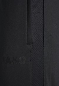 Närbild av svart JAKO sportklädseltyg med meshnätpanel och subtilt märkeslogotyp nära en vertikal söm.