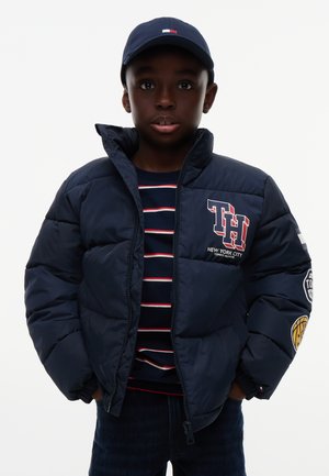 Dreng iført navy cap, navy dynejakke med logoer og navy stribet skjorte, stående med hænderne i jakkeærmerne mod hvid baggrund.