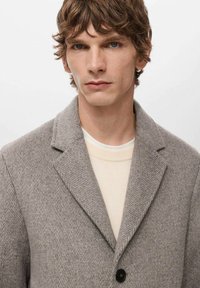 Grijze visgraatjas met een notched kraag, enkele zwarte knoopsluiting, gedragen over een licht crèmekleurige trui. Gladde woltextuur.