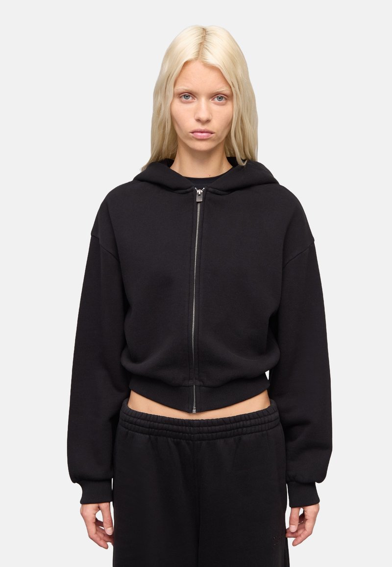 Sudadera corta negra con cremallera frontal, capucha grande y dobladillo acanalado. La tela parece suave con una textura lisa. Combinada con pantalones de chándal negros.