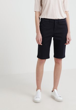 Persoon die zwarte knielange shorts, witte sneakers en een lichtbeige top met korte mouwen draagt, staand tegen een effen achtergrond.