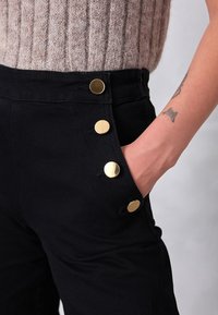 Czarne spodnie z denimu z wysokim stanem, złotymi guzikami i boczną kieszenią, zestawione z fakturowanym jasnym swetrem.