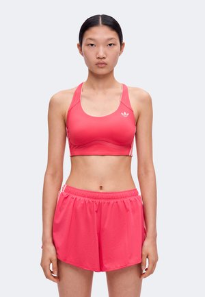 Jeune femme debout portant un soutien-gorge de sport rose et un short de sport rose assorti sur un fond clair.