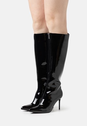 Bottes montantes en cuir verni noir brillant avec bouts pointus, fermeture éclair latérale et talons aiguilles portées sur des jambes nues sur un fond uni.