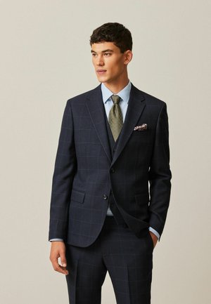 Chaqueta de traje a cuadros en azul marino, con forma ajustada, combinada con pantalones a juego. Lleva una camisa azul clara y una corbata verde estampada. El pañuelo de bolsillo añade un toque de detalle.