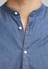 Camicia in denim blu con colletto alla mandarina, dotata di una lista di bottoni e due bottoni bianchi. Il tessuto ha una finitura liscia e leggermente strutturata.