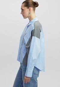 Camisa de botones azul claro con paneles laterales grises, un corte relajado y adornos decorativos plateados en la parte delantera. Jeans de mezclilla usados abajo.