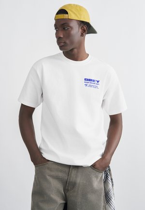 Νεαρός άνδρας που φοράει λευκό t-shirt με τη λέξη «OBEY», κίτρινο καπέλο με το ανάποδο φοράρισμα και γκρι παντελόνι με ένα καρό πουκάμισο που είναι βαλμένο στην τσέπη, κοιτάζει προς το πλάι.