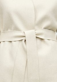 Μπεζ παλτό τύπου wrap με απαλό ύφασμα, ζώνη στη μέση με επίπεδη ζώνη και διακριτικές ραφές κατά μήκος των άκρων.