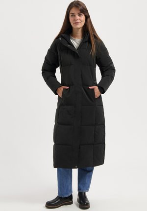 Frau in einem langen schwarzen gesteppte Mantel mit Händen in den Taschen, blauen Jeans und schwarzen Stiefeletten, die vor einem schlichten Hintergrund steht.