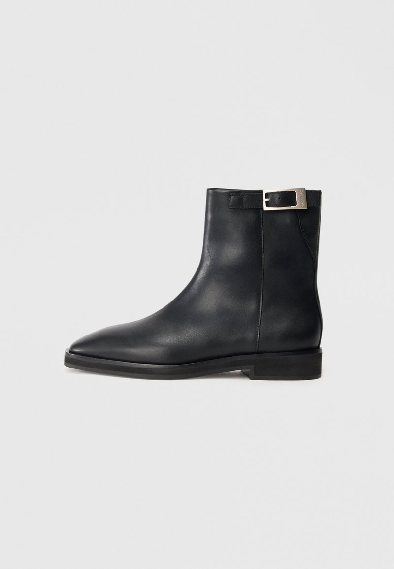 Bottine en cuir noir avec une bride à boucle argentée, bout pointu et petit talon carré sur un fond gris clair uni.