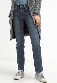 Vaqueros de denim azul oscuro con corte slim, ligero desgaste y diseño clásico de cinco bolsillos, combinados con zapatillas de deporte gris claro.