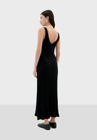 Zwarte geribbelde maxi-jurk met een diepe rug, voorzien van brede schouderbanden. Model draagt platte, donkergrijze schoenen en staat tegen een neutrale achtergrond.