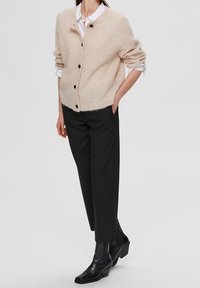 Cardigan lavorato a maglia beige con collo rotondo, caratterizzato da bottoni neri, indossato sopra una camicia bianca. Abbinato a pantaloni neri aderenti e stivali alla caviglia.