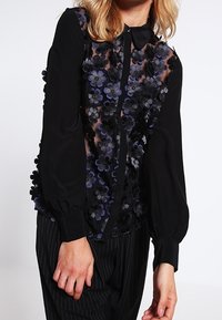 Svart blus med genomskinlig framdel, prydd med lager av tygblommor i mörkblått och svart. Har krage och långa puffärmar.