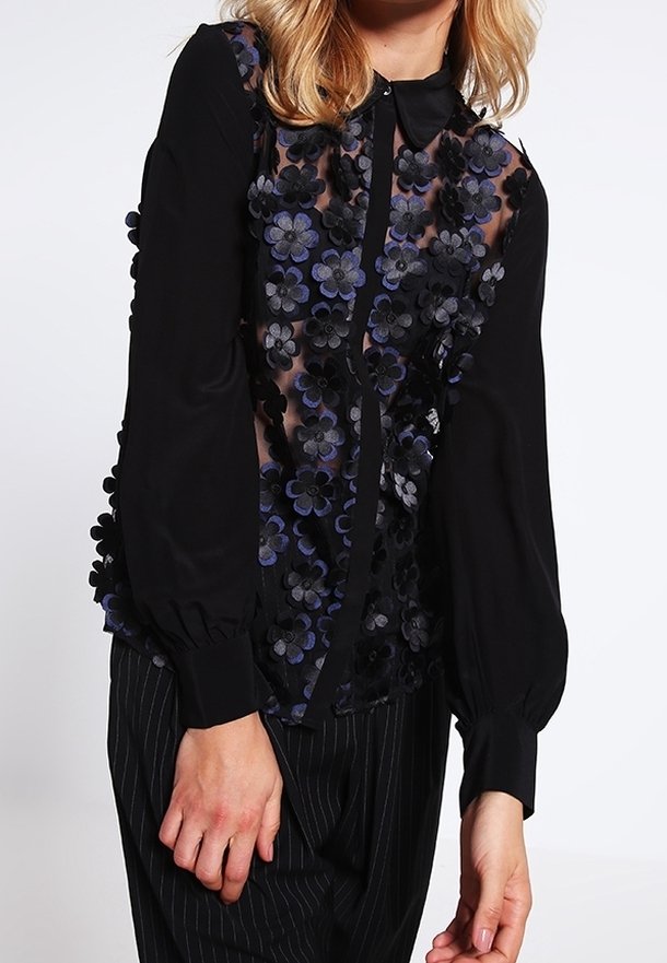Svart blus med genomskinlig framdel, prydd med lager av tygblommor i mörkblått och svart. Har krage och långa puffärmar.