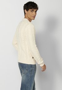 Suéter de punto trenzado color crema con mangas largas, puños y dobladillo acanalados, combinado con jeans azules. El suéter presenta patrones texturizados y un pequeño detalle de logo.