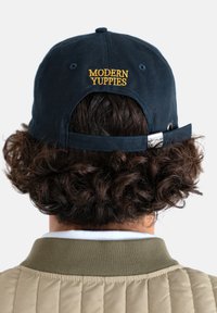 Tummansininen baseball-hattu, joka on valmistettu puuvillasta. Hatussa on keltainen brodeerattu teksti "MODERN YUPPIES" takana, säädettävä hihna ja metalliset osat.