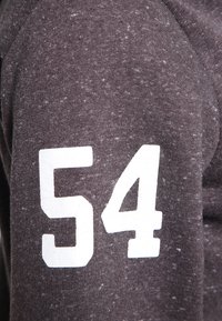 Tela texturizada de color gris oscuro con el número blanco "54" impreso en la manga. El material exhibe un patrón moteado, lo que indica un diseño informal.