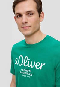 Groene katoenen t-shirt met witte tekst: "s.Oliver ELEVATED ESSENTIALS SINCE 1969." Korte mouwen, klassieke ronde hals, gladde textuur.