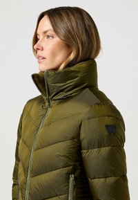 Oliwkowa kurtka puchowa z wysokim kołnierzem, zamkiem błyskawicznym i poziomym pikowaniem. Posiada czarną łatę z logo na ramieniu.