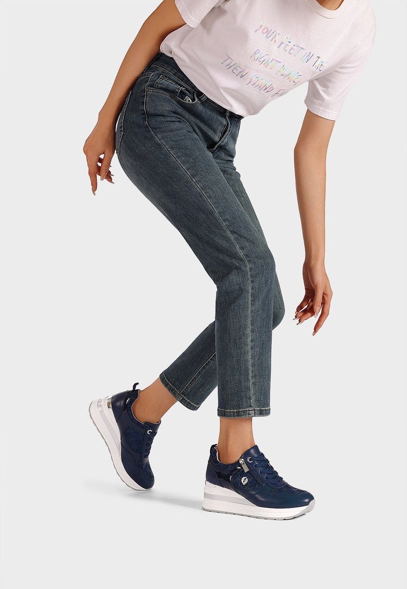 Femme portant des baskets compensées bleu foncé, un jean en denim bleu et un t-shirt blanc avec un texte coloré, posant contre un fond blanc.