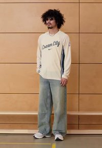 Camisola de manga comprida cor creme com texto "Cream City"; jeans soltos azul claro; ténis brancos; pessoa com cabelo encaracolado preto e óculos de sol.