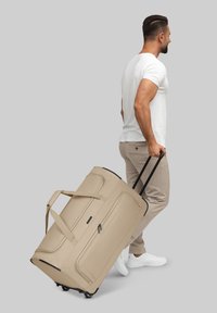 Beige Rolltasche mit zwei schwarzen Griffen, glatter Oberfläche, seitlicher Reißverschluss-Tasche und robusten Rädern für einfache Mobilität.