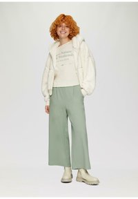 Witte, fluffy rits-hoodie over een beige, korte mouwen top met groene tekst; gecombineerd met losse, lichtgroene broeken en crème enkellaarsjes.