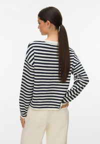 Femme avec une longue queue de cheval noire portant un pull rayé bleu marine et blanc ainsi qu'un pantalon crème, debout avec une main sur la hanche.