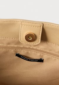 Beige taskeindvendigt med tekstureret stof, der har en lynlåslomme og et sort mærke med teksten "ANNA FIELD." Inkluderer en knaplukning med trykknap.