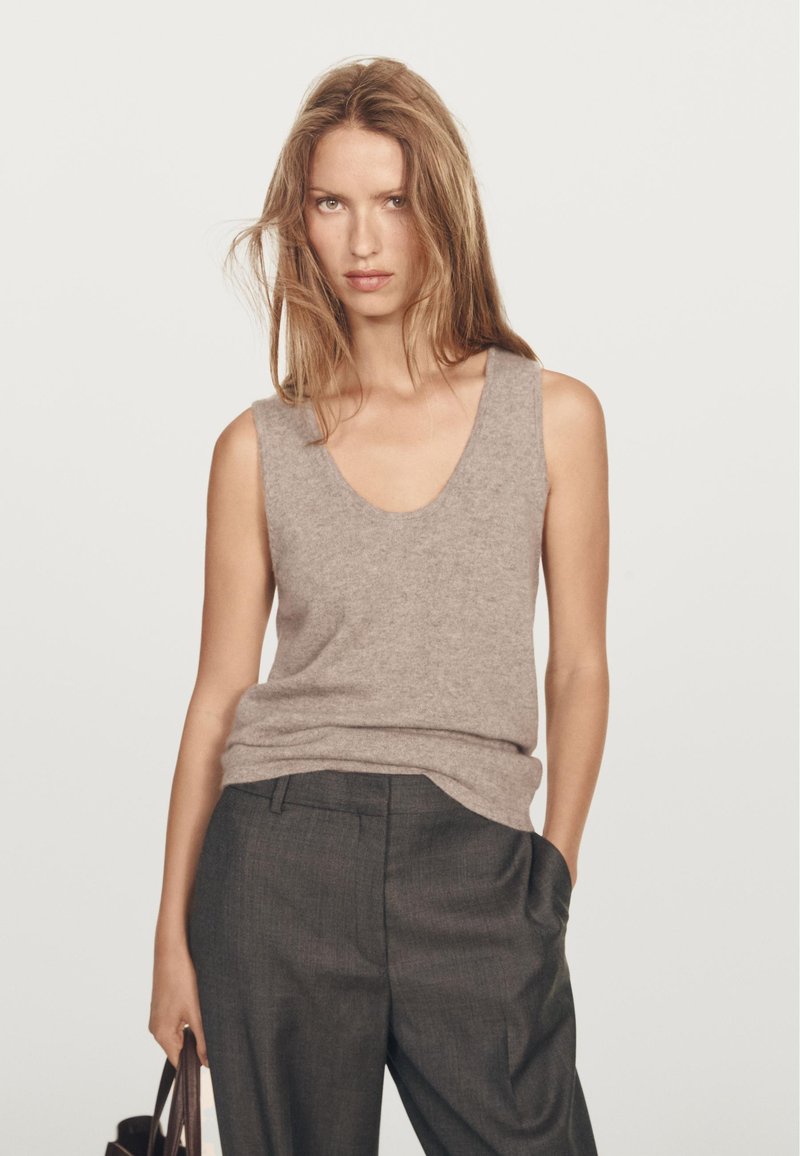 Žena s dlouhými světle hnědými vlasy, oblečená v béžovém bezrukávovém svetru a tmavě šedých kalhotách, držící hnědou kabelku a s jednou rukou v kapse.